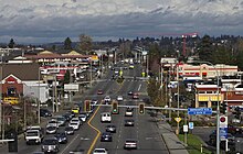 Una vista aérea de una calle suburbana con varios carriles de tráfico y semáforos en medio de centros comerciales y gasolineras.