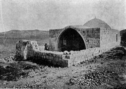 Uma fotografia em preto e branco que mostra um muro baixo de pedra envolvendo um pátio em frente a um prédio baixo com uma entrada através de um arco pontiagudo com uma pequena cúpula atrás