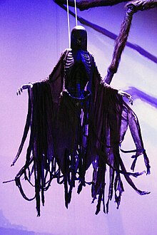 Farbfotografie in der Untersicht von einer dunklen Figur, dessen Gesicht nicht zu erkennen ist und die ihre Arme ausbreitet. An seinem skelettartigen Körper hängt ein schwarzer Umhang, der unten in mehreren Streifen zerrissen ist.