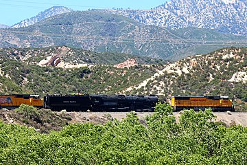 Two yellow diesel locomotives hauling an articulated steam locomotive, one at the front and the other at the back, through the mountains in the background