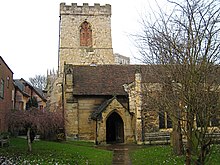 The western section of a stone church with a red tiled roof seen from the south, showing the porch and a battlemented tower