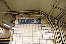 Foto der Wand einer U-Bahn-Station mit Rohrleitungen und einem Wegweiser-Schild, das mit einem Richtungspfeil den Weg zu den „BMT LINES“ weist.