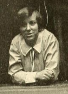 A young white woman, leaning forward and resting on her elbows, wearing a light-colored blouse with a wide tie collar