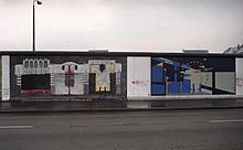Frontale Farbfotografie von der Berliner Mauer mit zwei Bildern. Das linke Bild zeigt eine abstrakte Maschine mit drei Stationen, die aus Tasten und Rohren bestehen. Das rechte Bild zeigt graue und blaue Rechtecke, die sich in der Mitte eines gelb-blauen Sternenkreises treffen.
