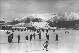 A male figure skater performing on a large frozen outdoor area with spectators and judges nearby on the ice. The background shows snow-covered mountains and a building.