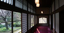 Un couloir de bois en enfilade, portes coulissantes en bois et papier sur la droite, fenêtres sur la gauche. tapis rouge au sol. À l’extérieur par les fenêtres, vue sur un jardin avec un cerisier.
