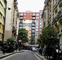 Foto colorida da fachada de um prédio de dez andares (imagem de fundo), vista de uma rua (primeiro plano) cercada por edifícios residenciais de quatro a cinco andares.