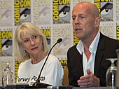 Bruce Willis berbicara di San Diego Comic-Con. Aktris Helen Mirren duduk di sebelah kanannya mengenakan kemeja putih dengan nama Harvey Pekar