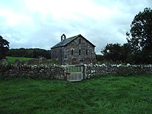 Eine kleine, einfache Steinkirche, vor der sich eine Steinmauer befindet. Auf dem fernen Giebel befindet sich eine Glocke mit einer einzelnen Glocke.