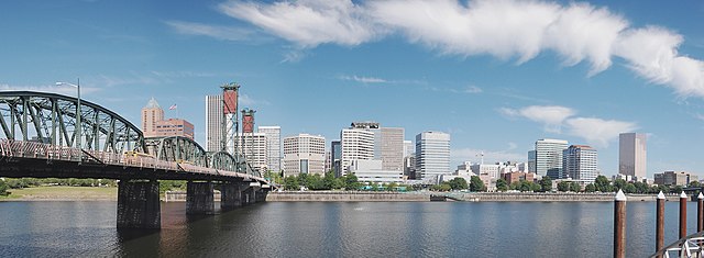 A city skyline at day, with a bridge and river in the forefront and the cityscape and partly cloudy sky in the background.