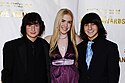 A photograph of three people, the two on either end being boys wearing black suits and the one in the middle being a girl wearing a purple dress