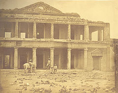 Braune Sepia-Fotografie von einem zerstörten Gebäude mit vier Menschen und einem Pferd davor. Das Gebäude besteht nur noch aus Säulen und Außenmauern mit Durchbrüchen.