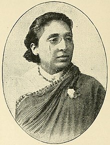 A middle-aged Indian woman wearing eyeglasses and a sari with a white blouse, and a flower pinned to the chest.