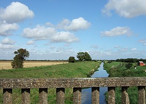 From the parapet of a lichen covered concrete bridge we are looking along the line of the navigation, which stretches dead straight to the horizon. It is well filled with placid water. There are wide, grass-covered embankments on each side, with wide grassed tracks on top. To the side are fields of ripened wheat, the nearest just harvested. A few oak trees stud the distant embankments. It is a glorious summer day with a deep blue Lincolnshire 'big sky', studded with small fluffy white clouds.