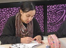 Jeune femme aux cheveux noirs ramenés en arrière, avec un erping dans la lèvre inférieure en train de dessiner sur la 2ème de couverture d'un manga