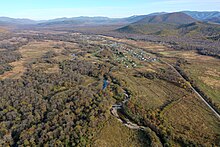 Photographie d'une vallée large avec de la végétation et des clairières, avec une petite rivière au centre et des monts en arrière-plan. Une route passe à travers la vallée à droite.