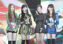 Blackpink standing in a row on stage in front of two microphones. Rose holds a bouquet of flowers while Jisoo holds a trophy.