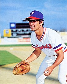 A man wearing a white baseball uniform with a blue cap on a field leaning forward, glove out, preparing to field a hit ball