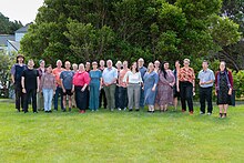 a group of about thirty people stand facing the camera smiling. They are standing on a lawn with trees in the background.