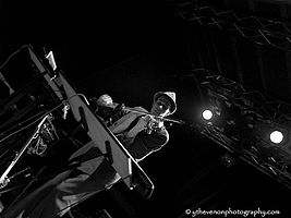 Diagonally tilted black-and-white photo of white male wearing a suit and hat, standing in front of a keyboard onstage, playing a violin.