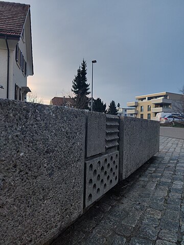 The concrete fountain which today is located at the intersection of Zihlstrasse and Gstaldenstrasse in Hinwil was designed by Max Vogt as part of the Hinwil railway station. It has since been moved away from the railway station and placed here. This photo is a close-up of the fountain's outer wall which is facing Gstaldenstrasse and features an interpretation of the Hinwil municipality's coat of arms using patterns to represent colours. The fountain is empty, presumably because water supply is turned off during winter