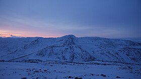 雪に覆われた山。視界の全体が暗い青色に見え、左側からは暗いピンク色の輝きが地平線から立ち上っている。これでも太陽の正中時の風景である。