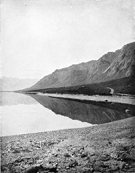 A rocky hill on the right, with trees on a flatter section at the base. A track goes through the trees to the rocky shore of a lake. The lake has a mirror image of the hills reflected in it.