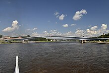 Photographie aérienne du pont piéton depuis un bateau sur la rivière. On distingue à droite le kremlin de Novgorod, et derrière le pont un trois-mâts.