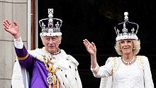 Charles and Camilla wearing their crowns and coronation robes waving from the balcony of Buckingham Palace