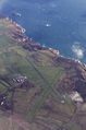 Widok z powietrza na lotnisko Alderney. Na zdjęciu widać trzy pasy startowe (jeden asfaltowy, dwa trawiaste), niewielką płytę lotniska oraz niewielki terminal. Lotnisko położone jest niedaleko brzegu, i jego okolica pokryta jest trawą i skałami. U góry zdjęcia widać skały gwałtownie opadające do morza, oraz niewielkie obłoki unoszące się nad ziemią.