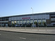 A three-storey building composed of glass windows and grey concrete stands across from a tarmac road and in front of a clear blue sky. A road passing horizontally in the foreground is divided by a pavement and metal barriers. The building's entrance appears central and is flanked by mirrored-glass windows and is marked by a red sign with the text BAE SYSTEMS.