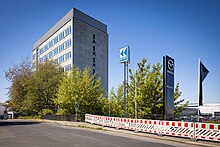 Foto eines mehrstöckigen, grauen Bürogebäudes, davor ein Stück Straße mit Baustellenabsperrung, einem Mazdawerbepfosten, einer dunklen Fahne und einem Werbewürfel mit der Aufschrift "Karl Köhler".