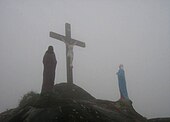 Foto av en Golgata med et kors og to statuer rundt den, på toppen av et fjell innhyllet i tåke.