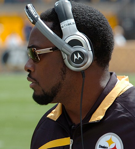 Mike Tomlin in profile on a football sideline wearing a black and gold Pittsburgh Steelers jacket, Motorola headset and sunglasses.