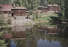 Foto von mehreren alt wirkenden Holzhäusern, im Vordergrund ein Gewässer