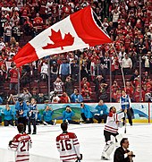 Ein helmloser Eishockeytorwart, der eine große kanadische Flagge an der Stange über dem Kopf trägt, während Teamkollegen, Zuschauer und Medien zuschauen. Er trägt ein weißes und rotes Trikot mit weißen Polstern.