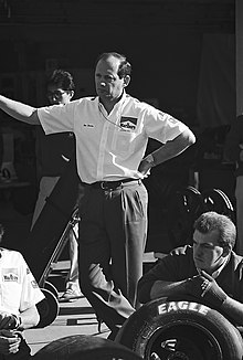 Photographie en noir et blanc d'un homme debout, croisant ses jambes, vu de trois-quarts, regardant à sa droite.
