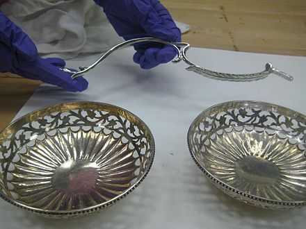 A tarnished and unpolished silver bowl (left) and a polished silver piece (right). There is a visible difference in cleanliness and color. The piece held above the two bowls has also been polished. (These pieces are part of an 18th-century silver epergne in the collection of the Indianapolis Museum of Art).