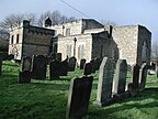 Im Hintergrund ist ein kleines Kirchengebäude aus Stein zu sehen.  Im Vordergrund ist ein Friedhof.