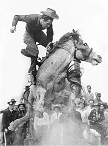 Um homem vestido em estilo cowboy está no ar, acima das costas de um cavalo, pulando em uma nuvem de poeira