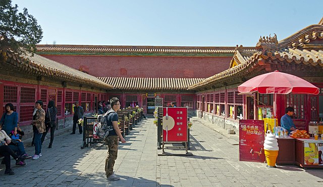 An area enclosed on three sides by red walls in traditional Chinese architectural style. All have screened windows and entrances; behind the rear wall is an even higher one. In the middle are tables, and at the right foreground is an ice cream cart and a red umbrella