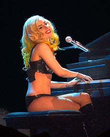 Right profile of a smiling, young blond woman with yellow streaks in her hair; she wears a black bikini, fishnet leggings and black leather boots and looks to her right while playing a black piano