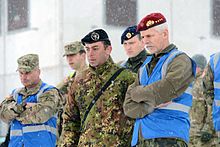 An older man with a gray beard, red beret, and olive green military suit.