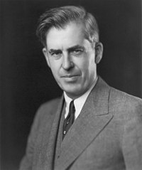 Head and shoulders of man about fifty with upswept hair, wearing a gray suit and dark tie