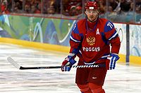 An ice hockey player is standing while slightly turned to his left. He has short dark hair and is not wearing a helmet. He is wearing a blue uniform with a large orange bird with an ice hockey stick on his chest.