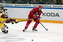 In der Mitte des Bildes ein Hockeyspieler in roter Kleidung zu Fuß;  ganz links ein anderer, überwiegend gelb gekleidet, beugte sich sehr vor und wollte den ersten mit seinem Stock behindern.