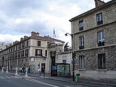 Photographie de l'entrée d'un hôpital, aux longs bâtiments grisâtres de deux étages depuis une rue.