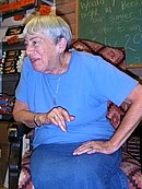 A photograph of a woman sitting in a chair, leaning on its armrest with her right arm, and looking over the armrest while wearing a blue shirt