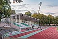 Tribüne mit grün-weiß-gestreiften Sitzbänken, von denen ein Stadion mit außenliegenden roten Laufbahnen beobachtet werden kann. Im Hintergrund Bäume und eine Beleuchtungsanlage