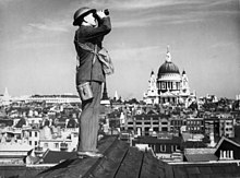 Un homme regarde le ciel avec des jumelles alors qu'il se trouve sur l'arête d'un toit. Une grande cathédrale se trouve à l'arrière-plan au milieu d'un paysage urbain.